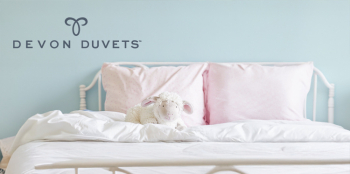 Neatly made bed with pink pillows and a soft toy lamb, under the Devon Duvets logo.