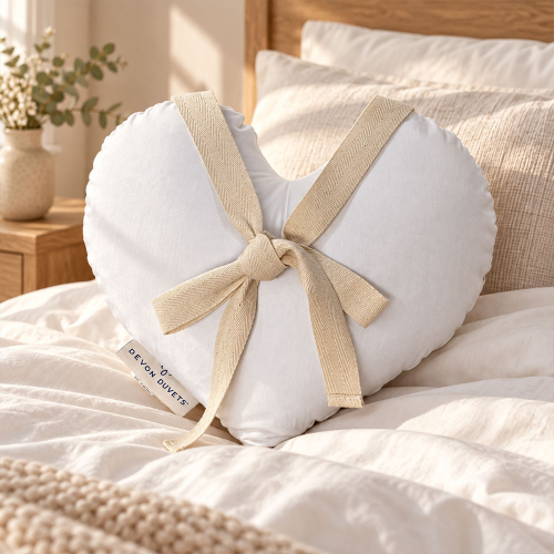 White heart-shaped Devon Duvets cushion with cream ribbon ties resting on a bed.