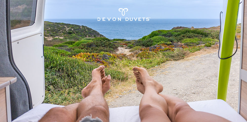 Close-up of a couple's feet on a plush Devon Duvets mattress topper, with serene beach and sea backdrop, symbolising travel comfort.
