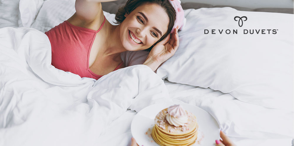 A smiling woman sitting in bed, with a stack of fluffy pancakes. She looks happy and relaxed as she enjoys her breakfast in bed.