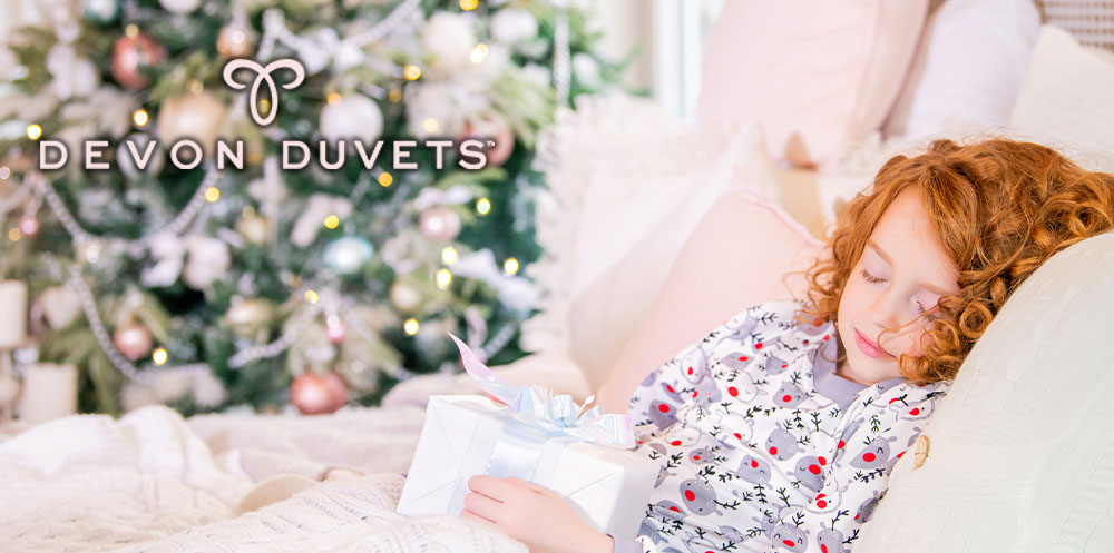 Young girl reading a festive storybook in bed, snugly wrapped in a Devon Duvets wool duvet, symbolizing a peaceful and comfortable bedtime routine on Christmas Eve.