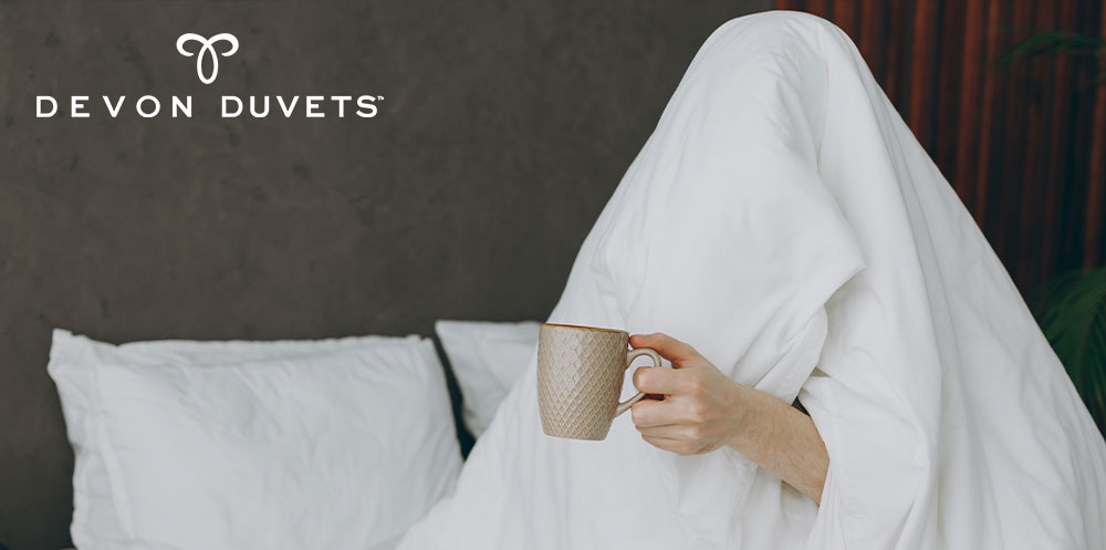 Person relaxing under a Devon Duvets wool duvet, holding a cup of coffee, symbolizing the balance between daytime caffeine consumption and restful sleep.