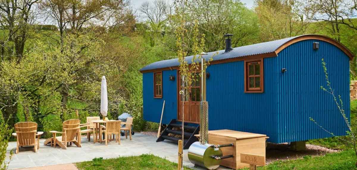 Charming blue shepherd’s hut with outdoor seating, set in a green countryside landscape.