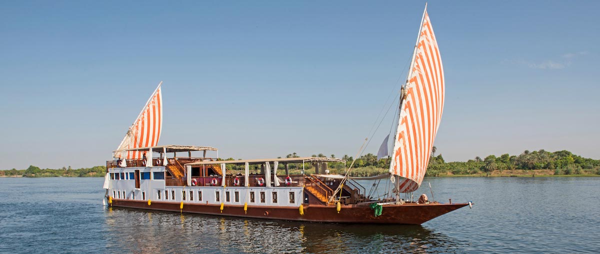 Classic wooden riverboat with striped sails cruising on calm water under a clear blue sky.